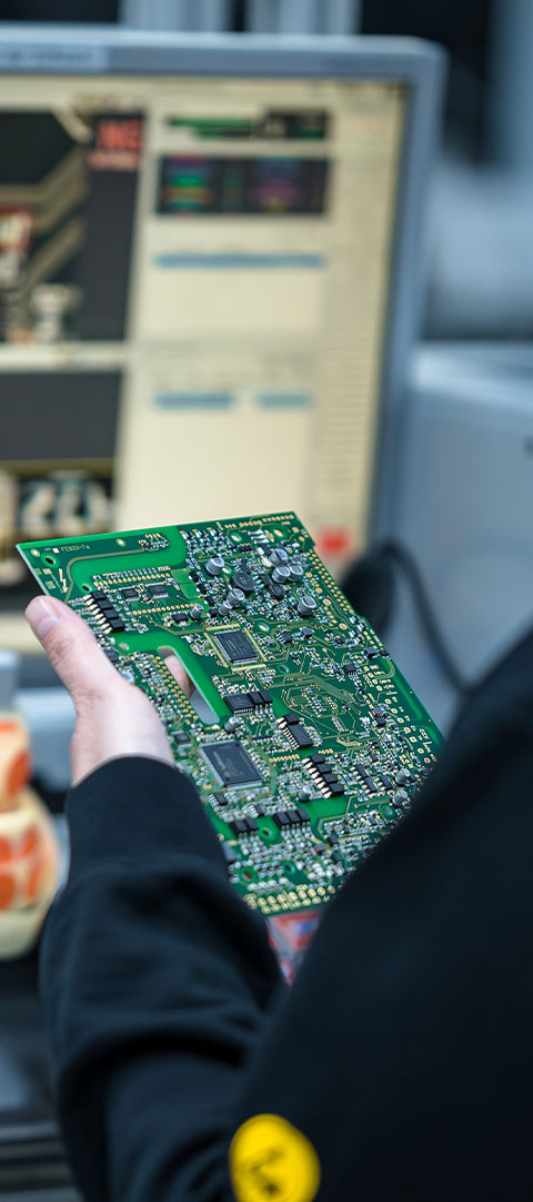 Hand hält eine bestückte Leiterplatte vor Bildschirm mit technischer Prüfsoftware
